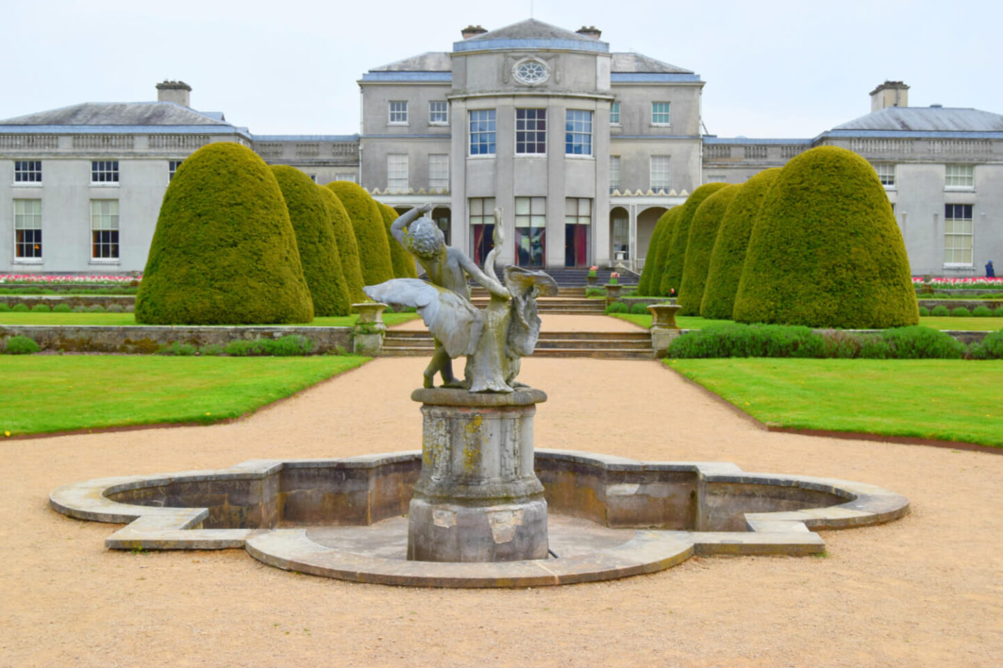 A view of a majestic fountain and a path way edged with yew trees leading up to Shugborough hall.