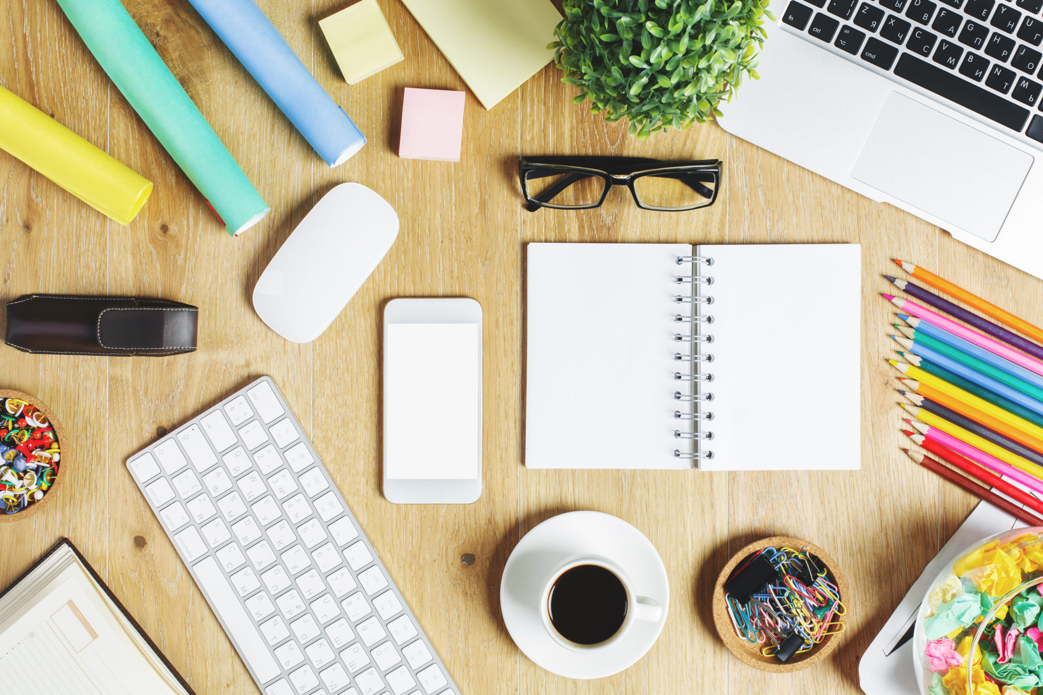 Looking at the top of a desk which has a keyboard, notebook, a mug of coffee, pencils and some glasses scattered around.