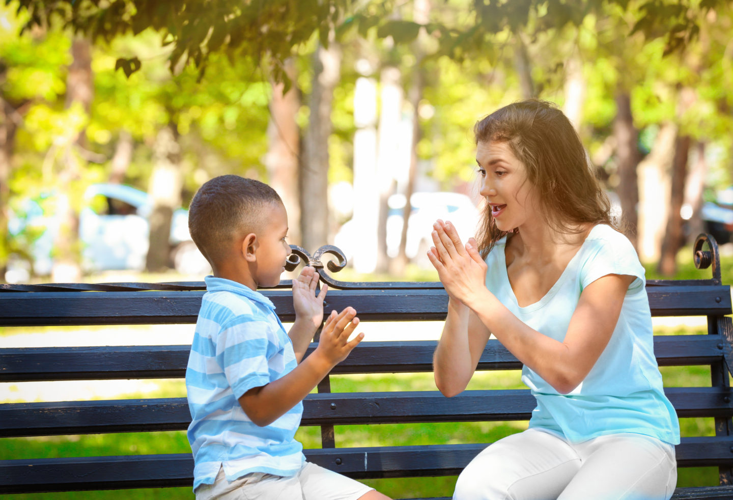 A foster carer sitting on a park bench playing a clapping game with a young boy.