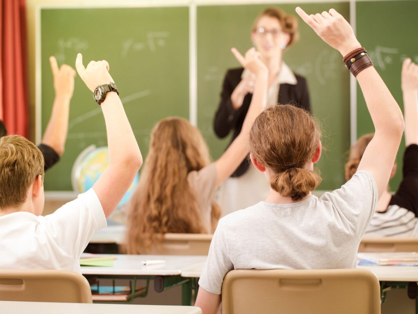 A classroom with high school children sat at their desks. They are holding their arms up in the air to answer a question.