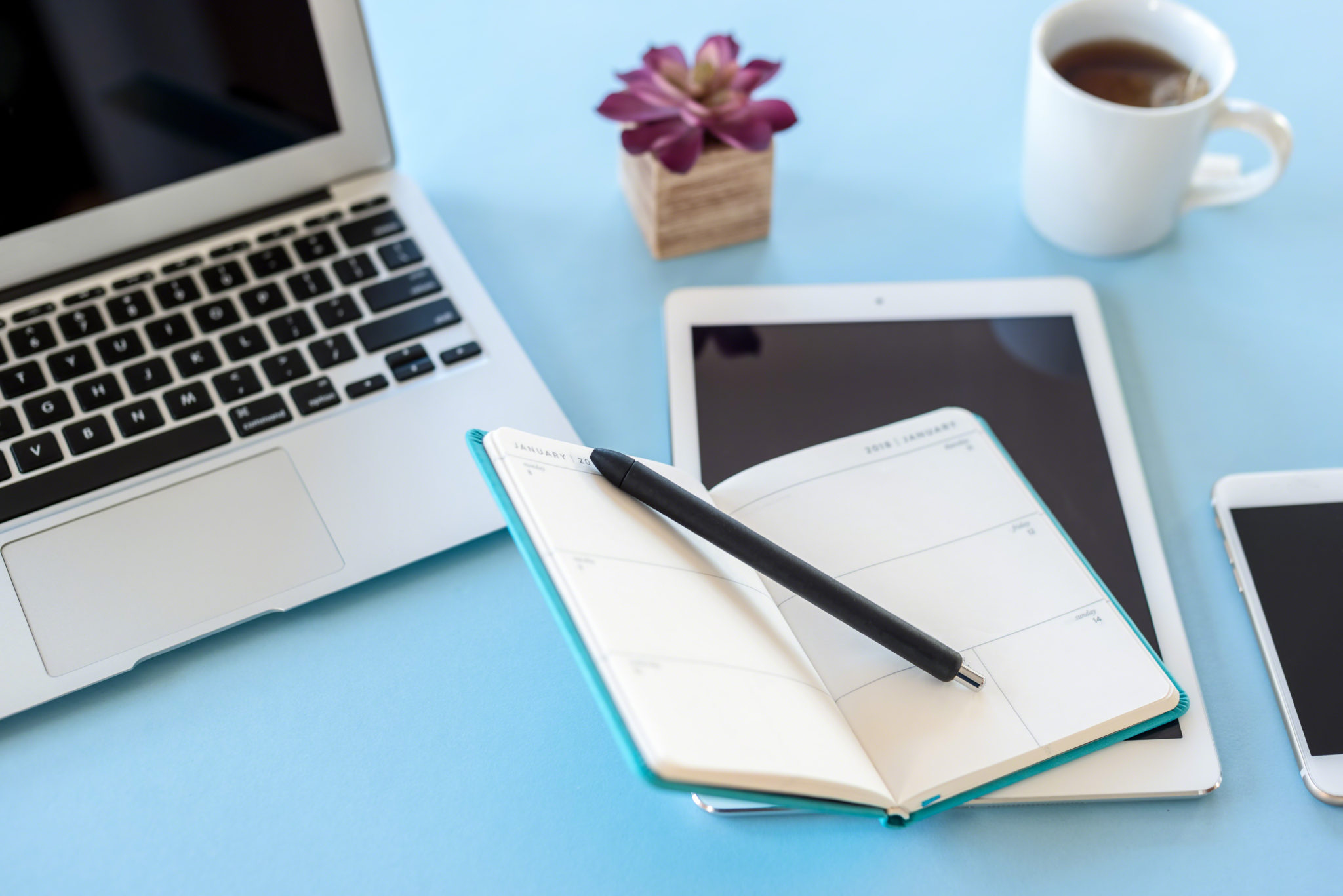 A bloggers desk with a laptop, ipad, phone, notebook and a cup of tea.