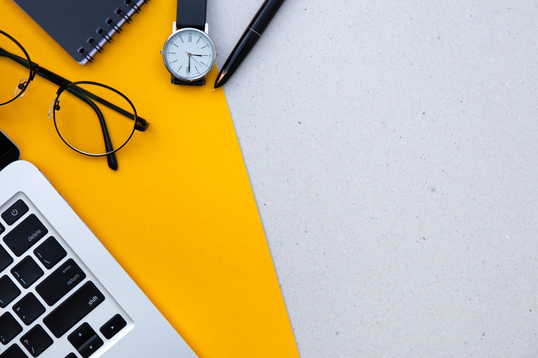A flatlay of a desk table. A keyboard, glasses, mouse, watch and en on a vibrant yellow background.