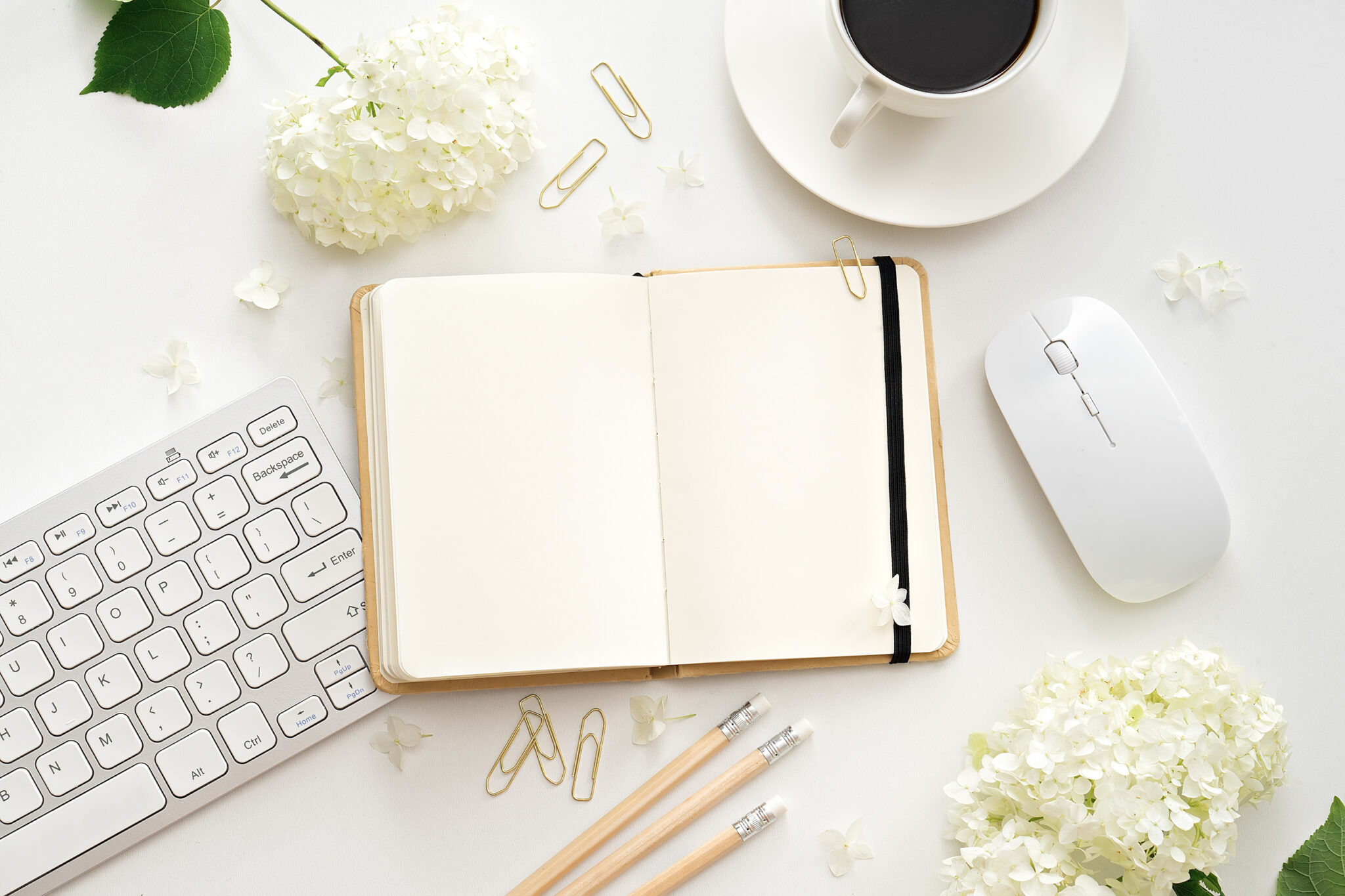 A spring themed cream desk flat lay. A desk top with an apple keyboard, notebook, fresh flowers and a cup of coffee.