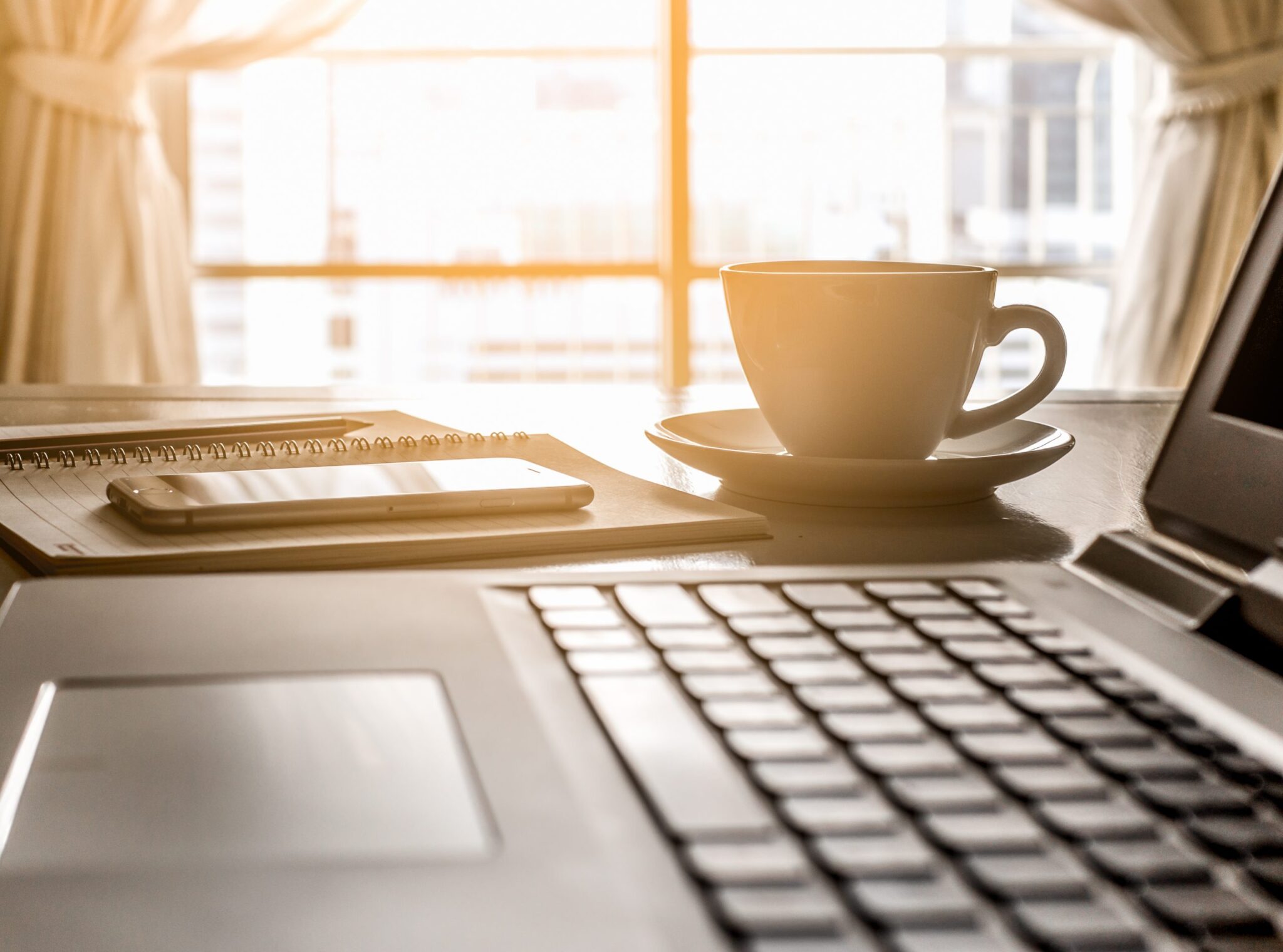 A desk with a laptop, cup, notebook and phone on top