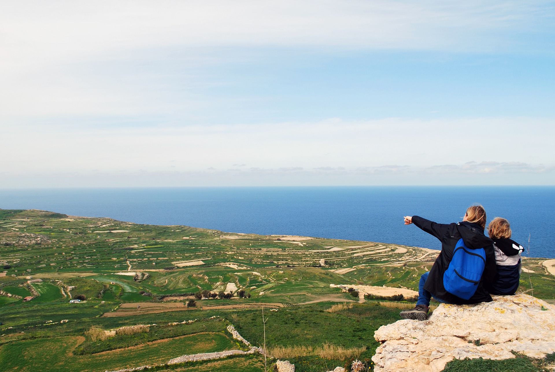 A mother and son sitting on a hill looking across green fields and the sea in the distance.