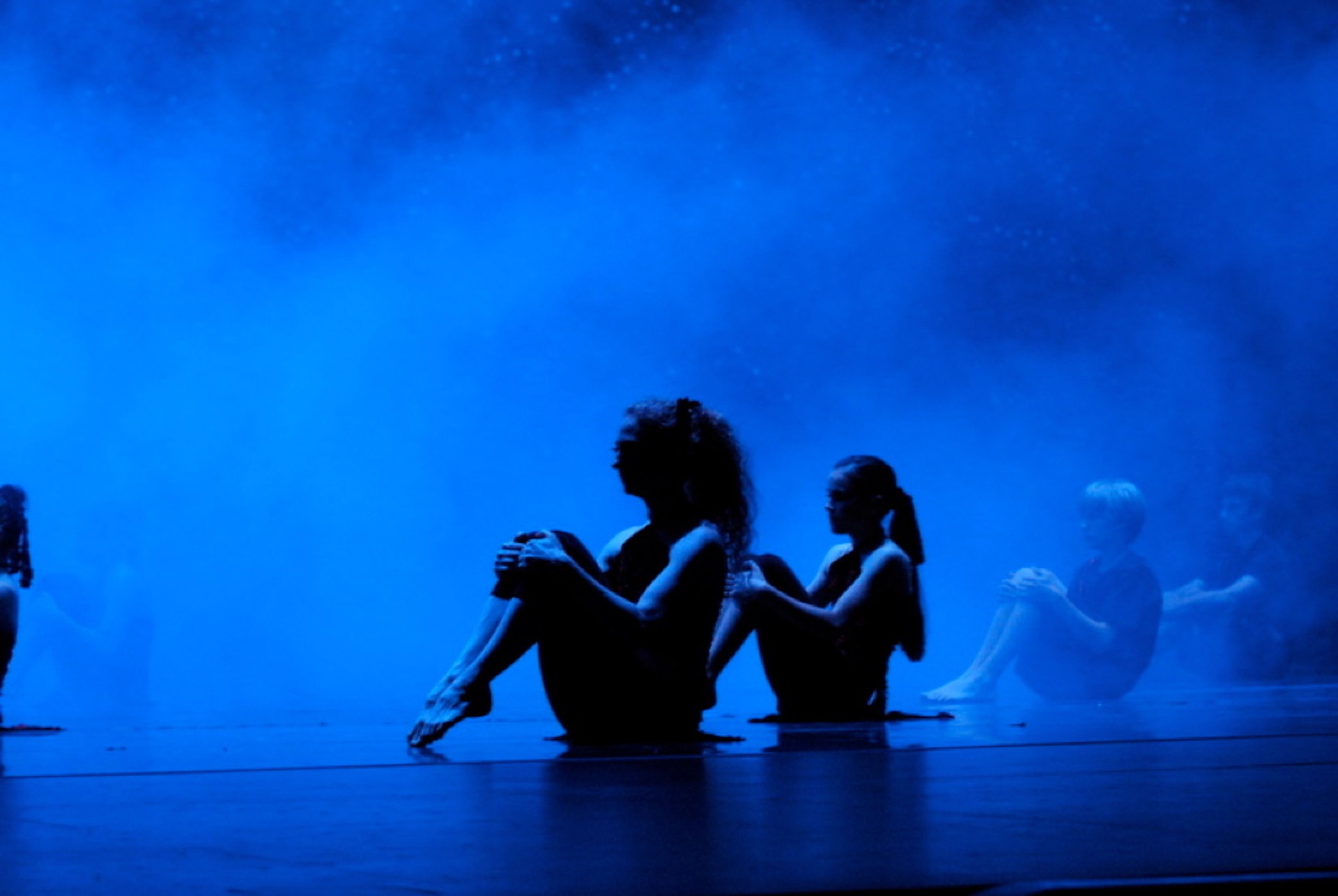 CHildren sitting down on a stage performing in front of a blue misty background.