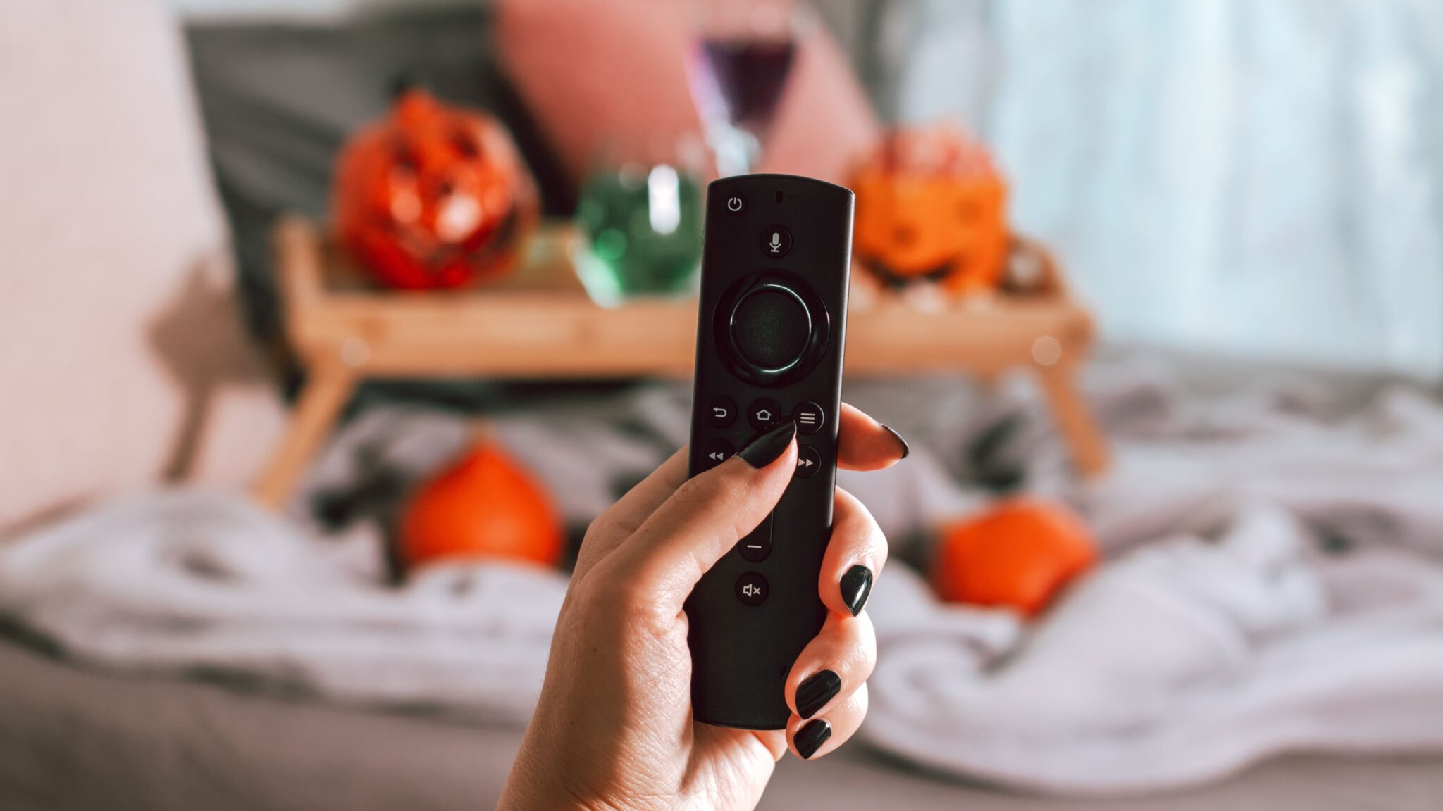 A lady's hand holding a TV remote with black painted nails and pumpkins in the background of the room.