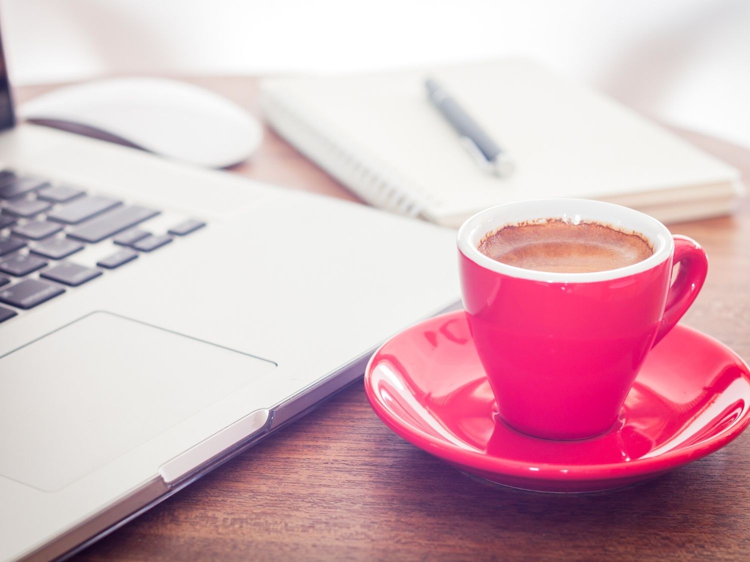A coffee in a pink cup and saucer on a desk top next to a laptop.