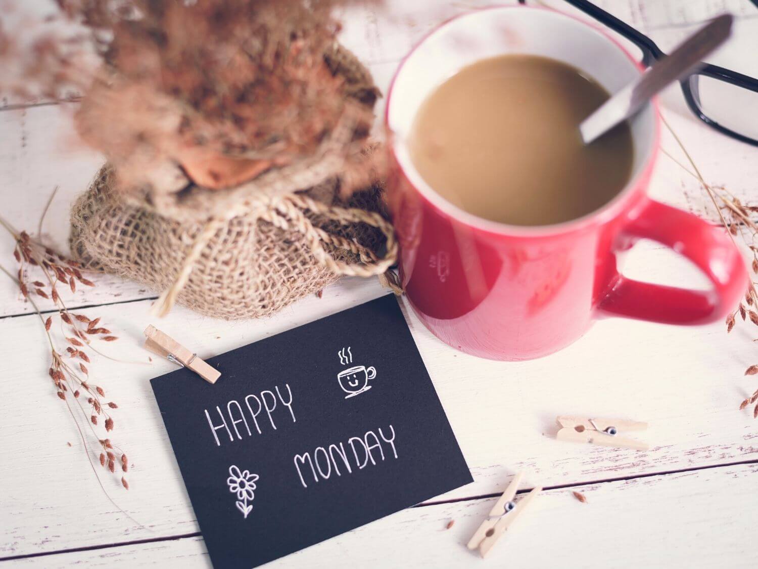 A mug of milky tea in a red mug next to a dried flower arrangement and a Happy Monday note.