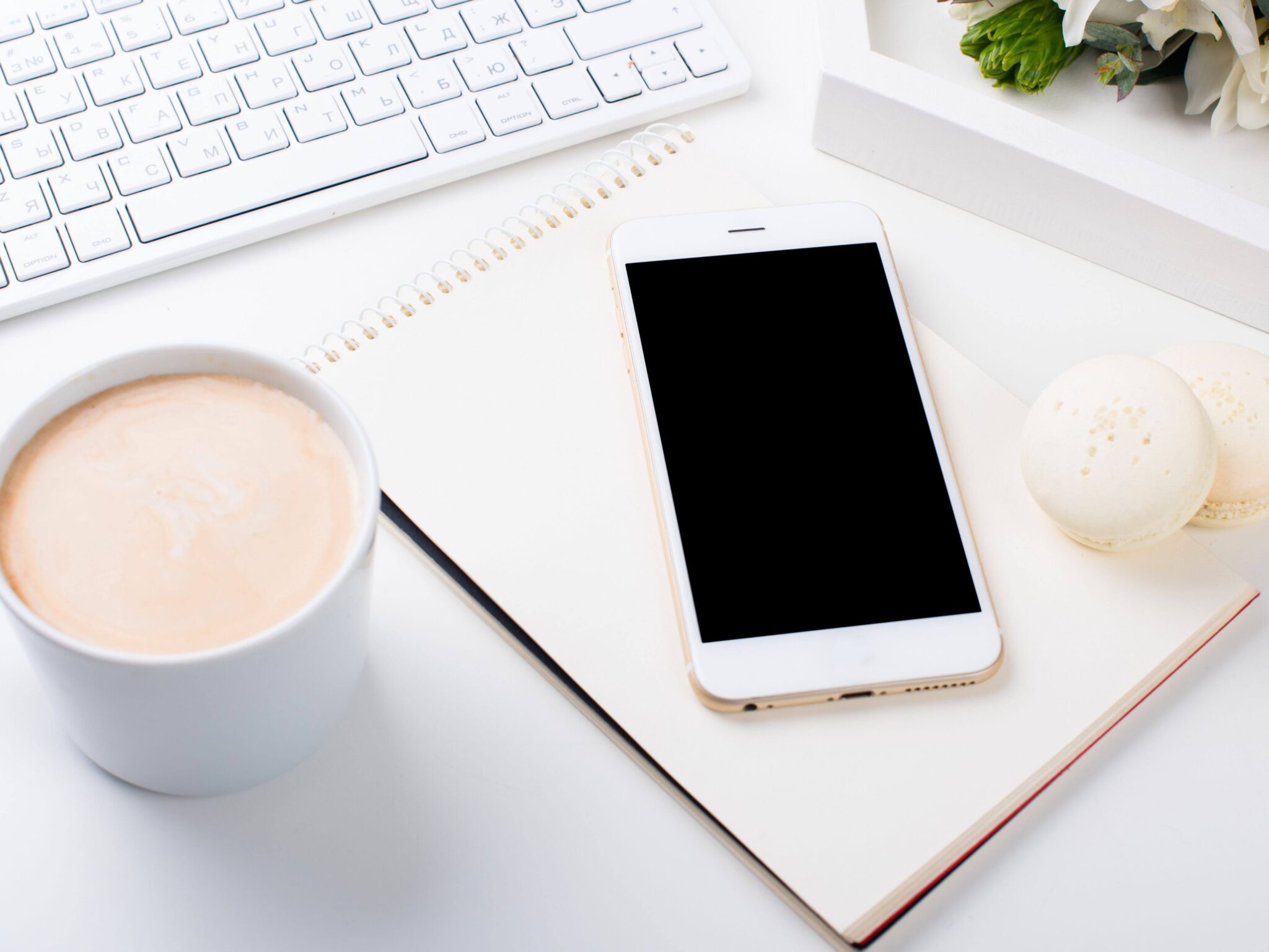A blogger's desk with an apple keyboard, a milky coffee, a notebook, a phone and a white macaroon cake.