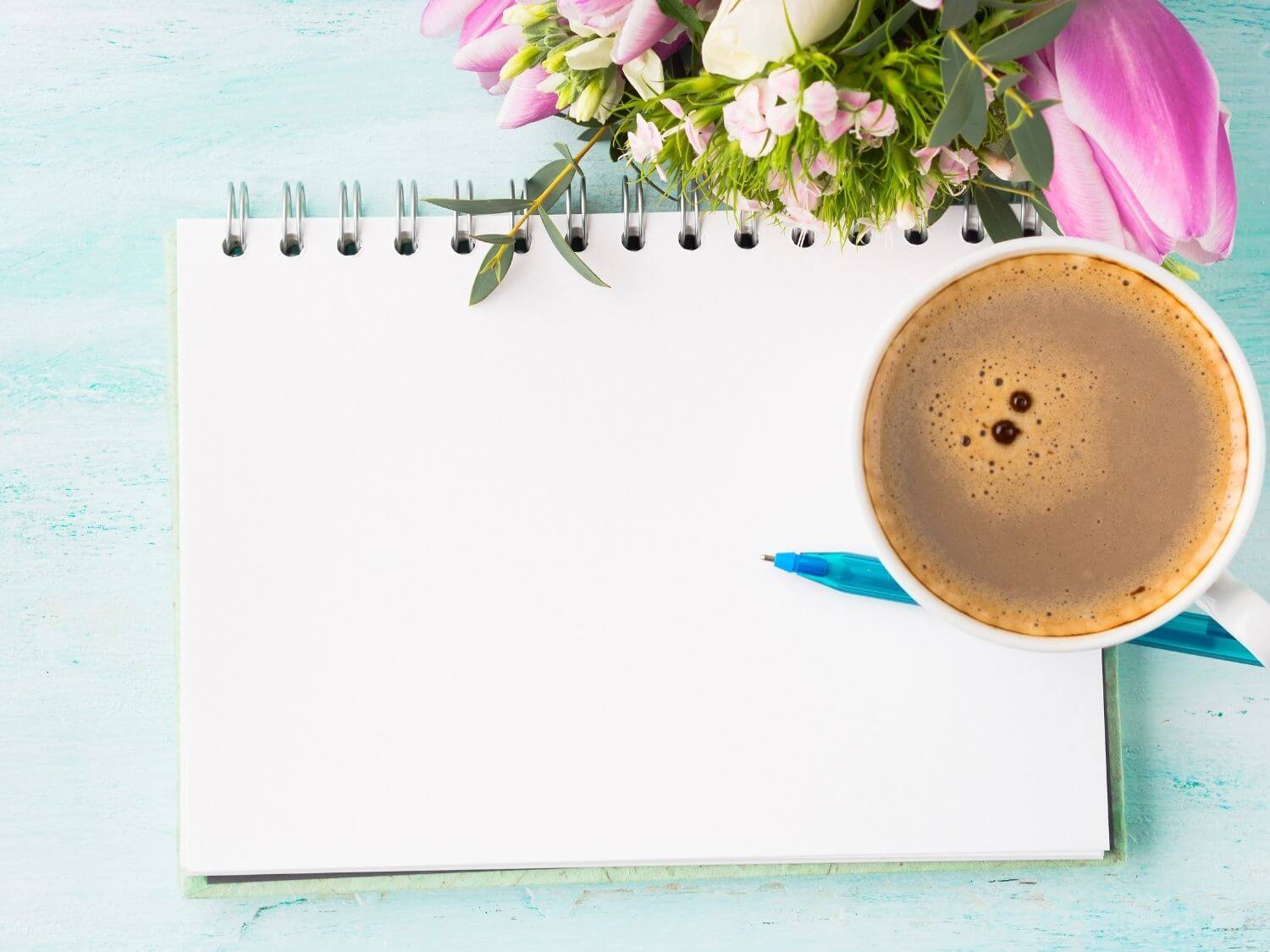 A morning coffee next to a notepad, a pen and a bunch of pink and white flowers on a desk.