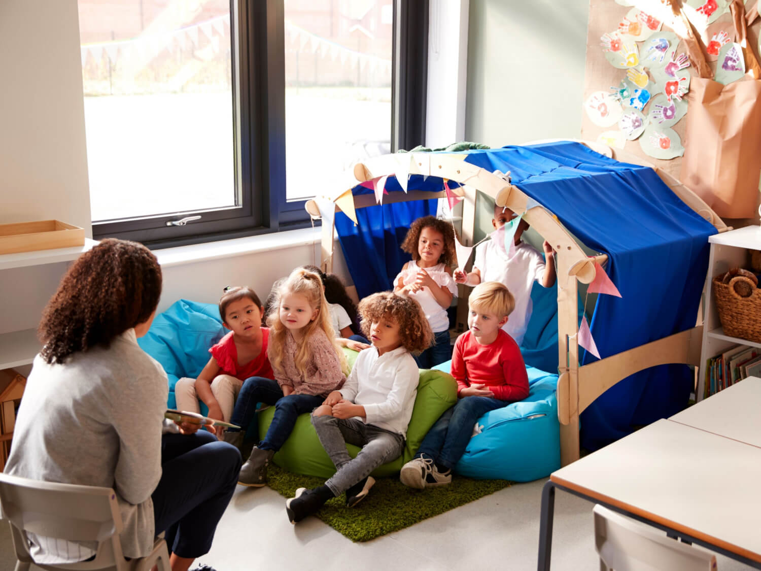 An infants school teacher reading a story to the children. They are sitting down in a little camp area, with a canopy above them.