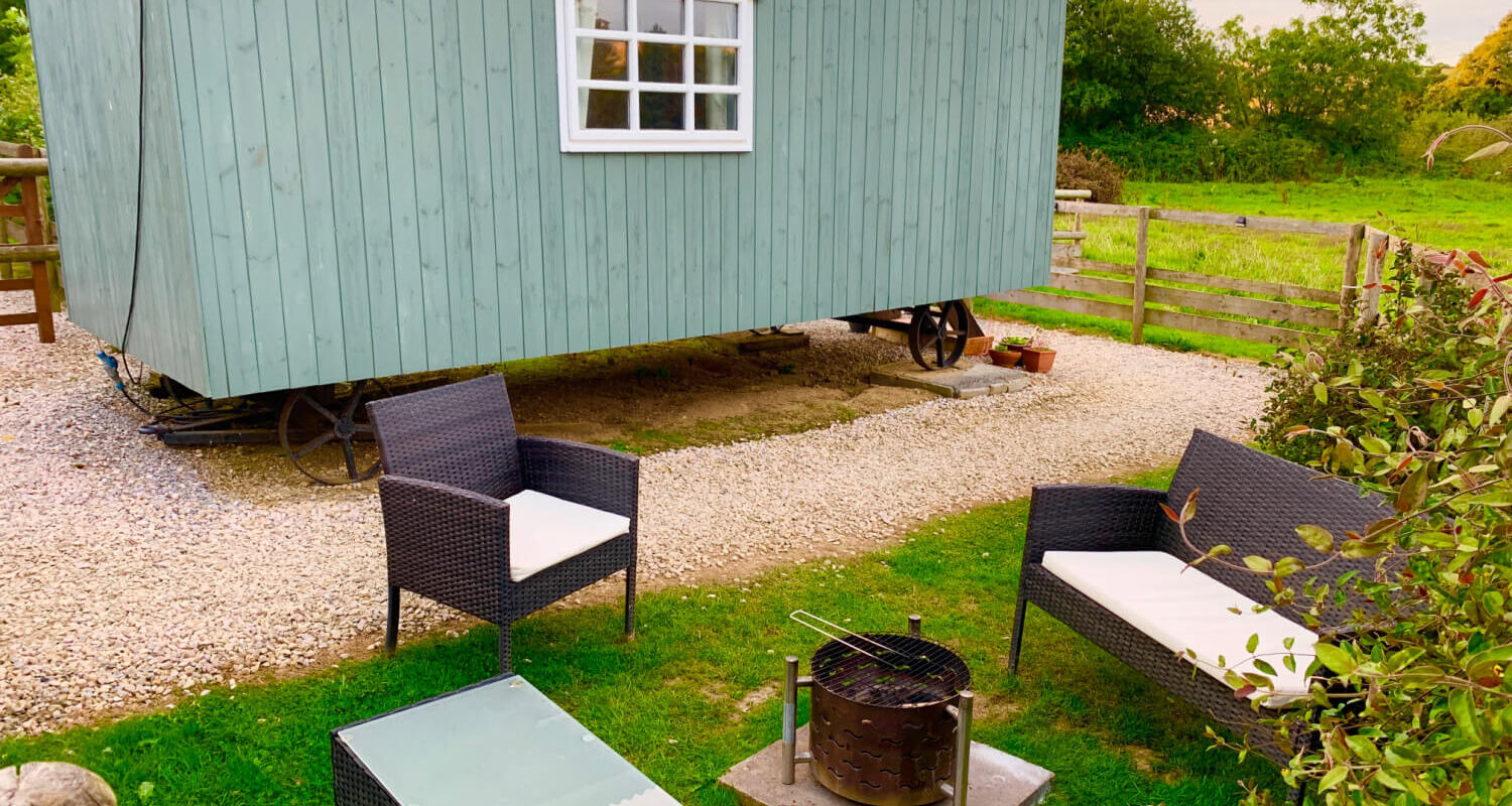 A teal coloured Sheperd's hut with a patio area and rattan furniture with a firepit at Hemsford yurt camp, Devon