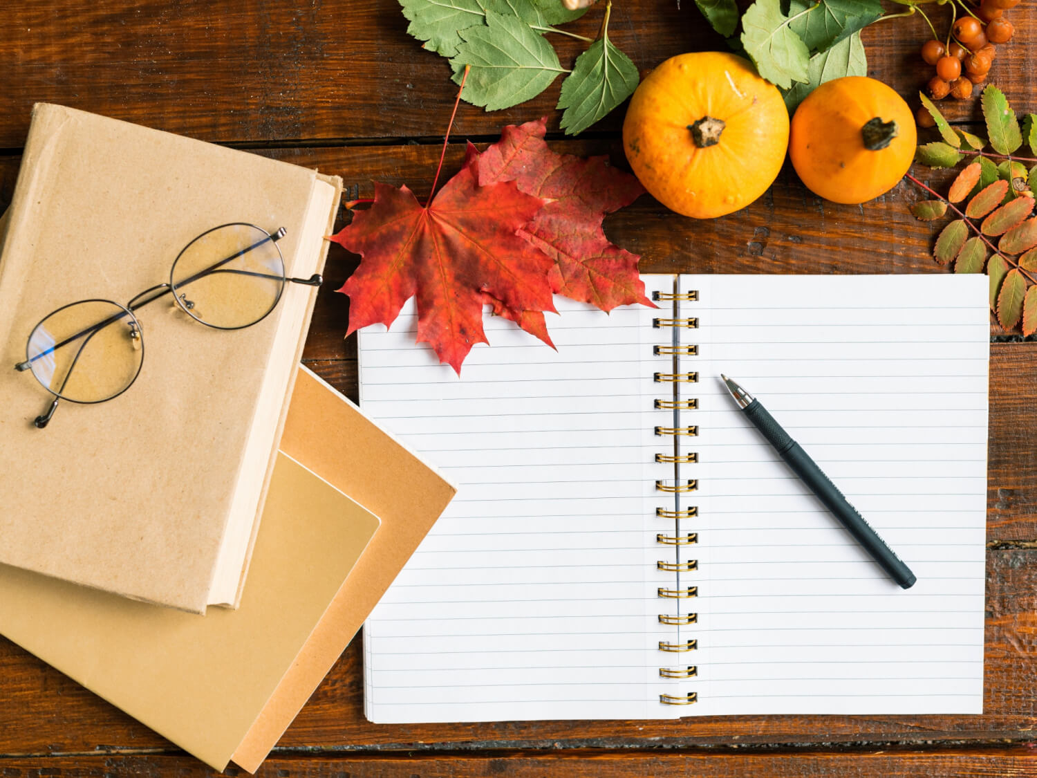 An open notebook on a desk with a pair of glasses and autumnal leaves and small pumpkins.