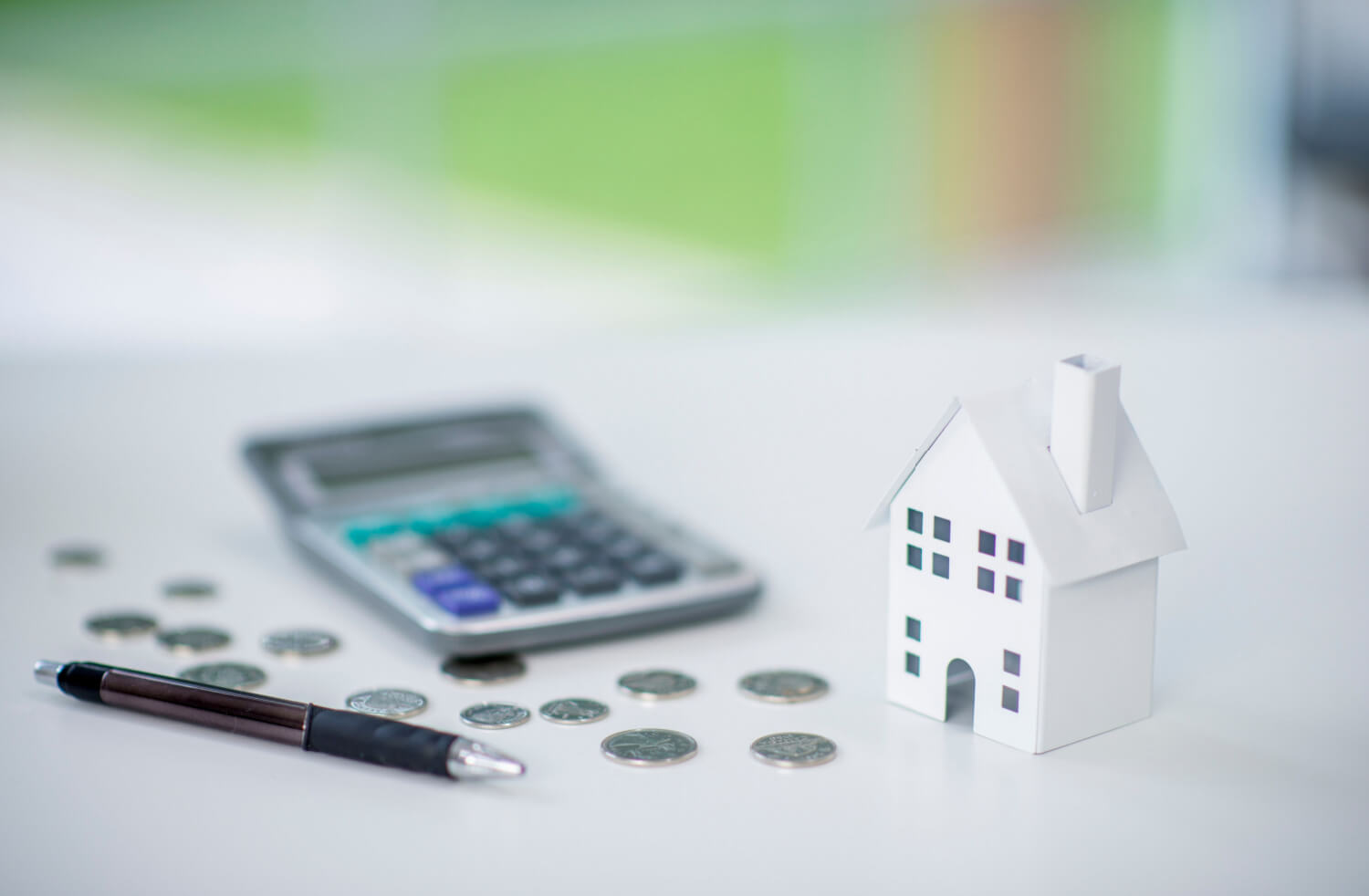 A white model house on a desk with a calculator, some coins and a pen.