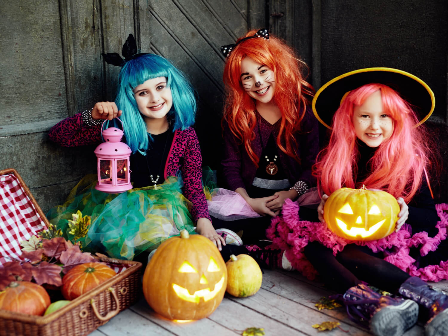 Three girls wearing coloured wigs and witches hats, sitting on a porch with pumpkins.