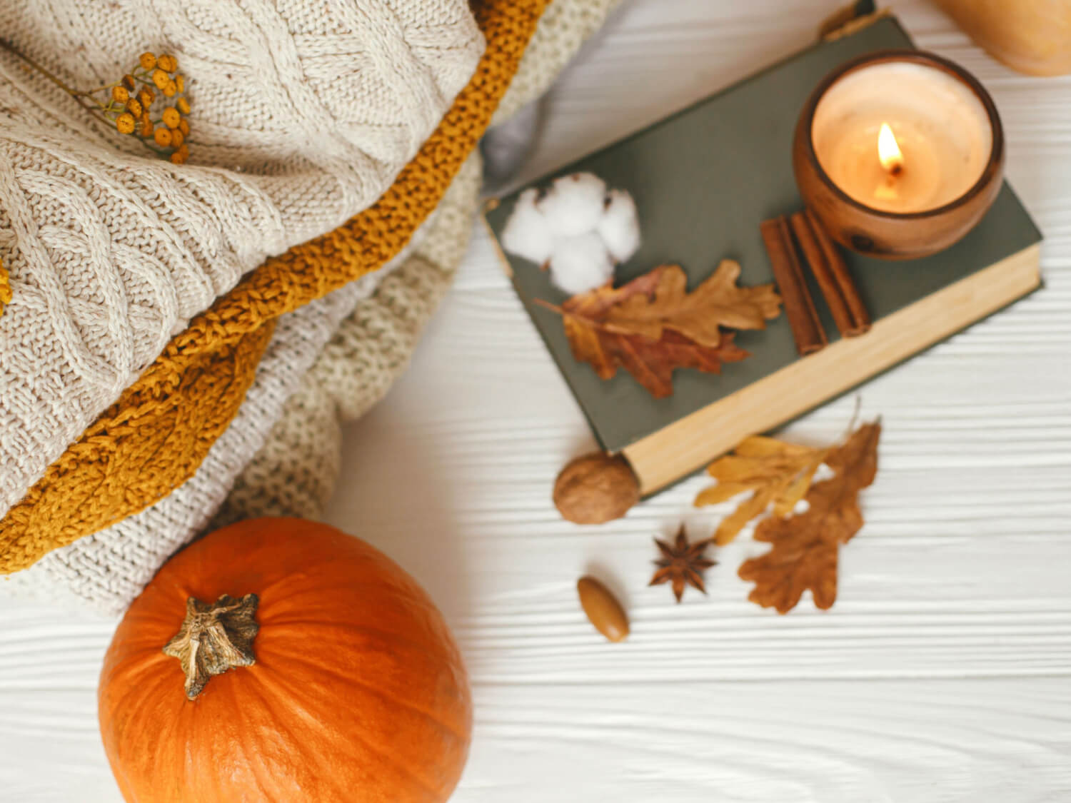 A white desk top with a orange pumpin, a notebook, some red leaves, a cozy blanket and a burning candle.