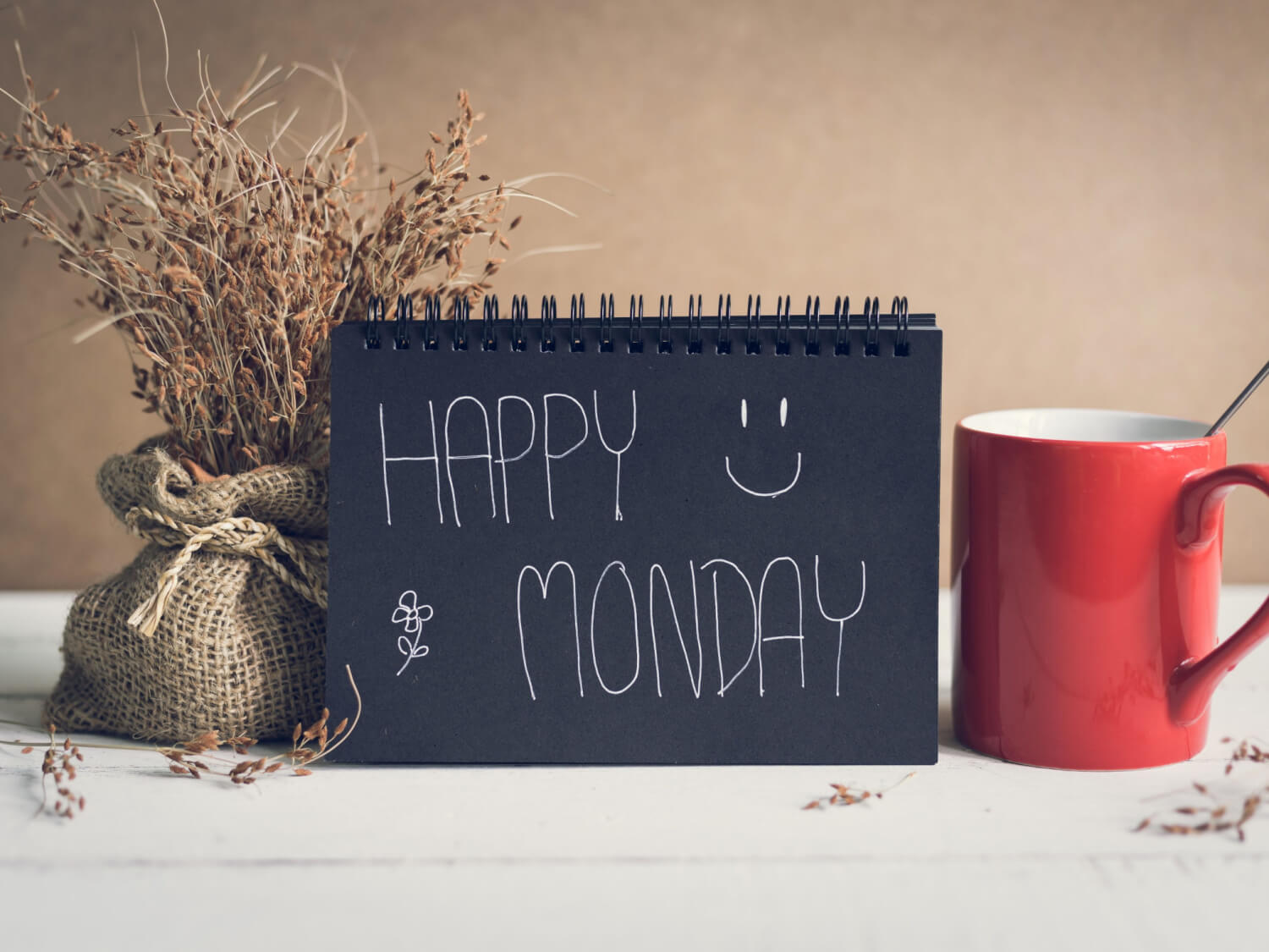 A black notebook with the words 'Happy monday' written on it. Next to the notebook is a red mug of tea and a dried flower arrangement in a jute bag.