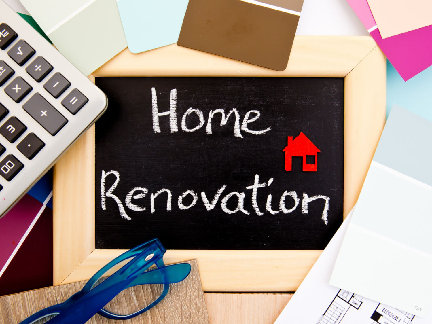 A chalkboard on a desk with the words Home Renovation written in chalk. There are also paint swatches and a calculator to the side.