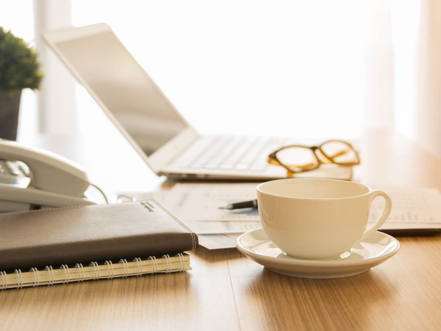 A desk beside a window with a mug of tea, an open laptop, notebooks and a pair of brown framed glasses