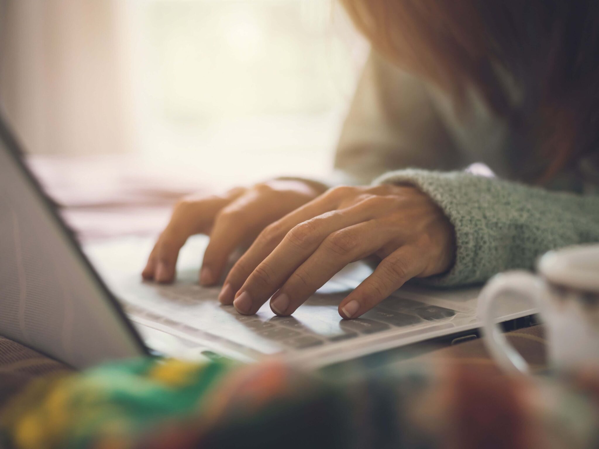 A lady typing on her laptop sitting in front of a window.