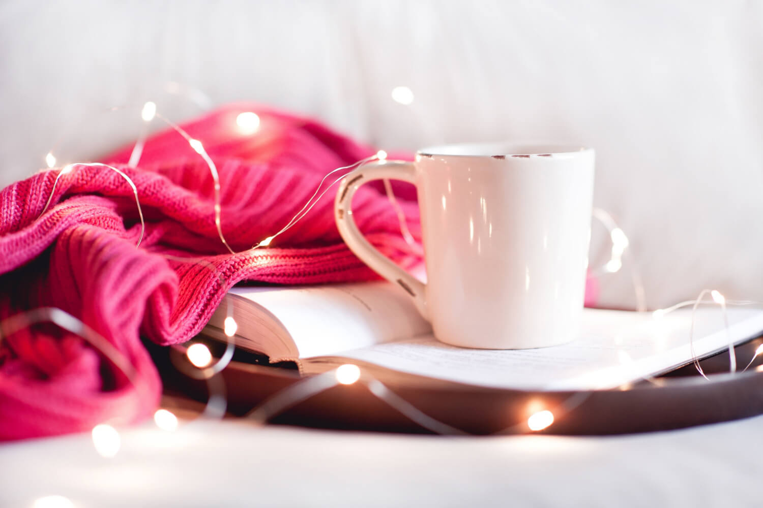 A mog of tea in a white mug which is by an open book, a pink blanket and warm white fairy lights.