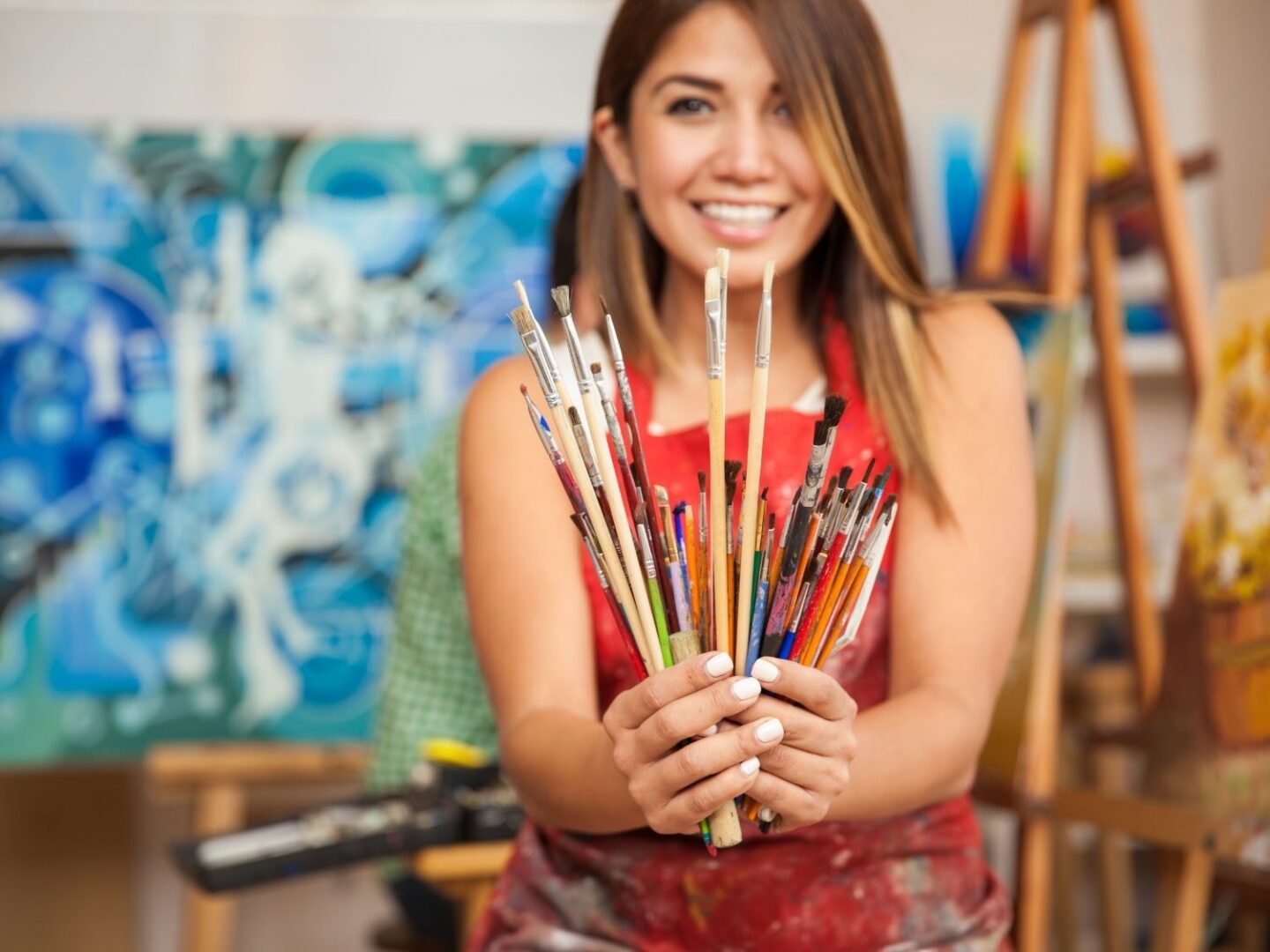 A lady holding paintbrushes with some of her artwork in the background.