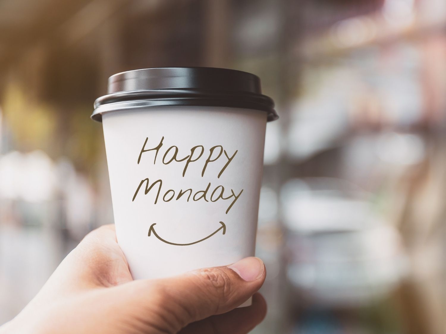 A hand holding out a travel cup with the words "Happy Monday" and a smile drawn beneath.