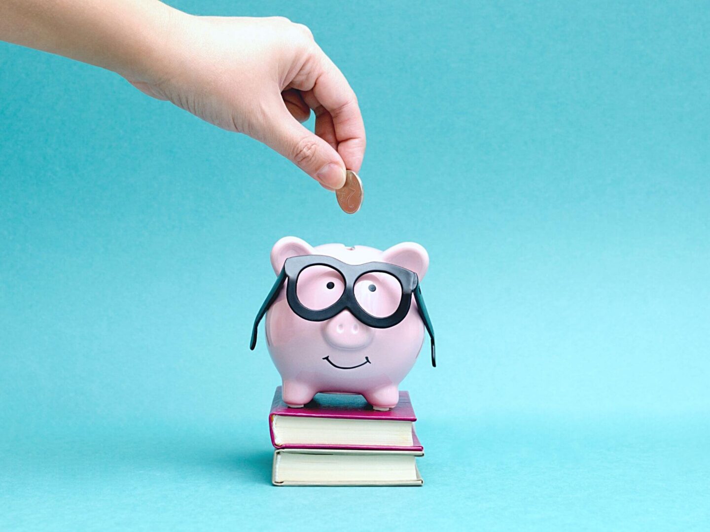 A hand putting a coin into a pink piggy money bank wearing glasses.