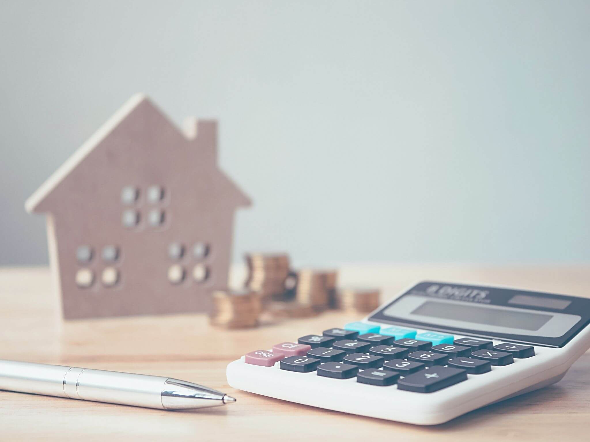 A wooden model house with a calculator and piles of coins.