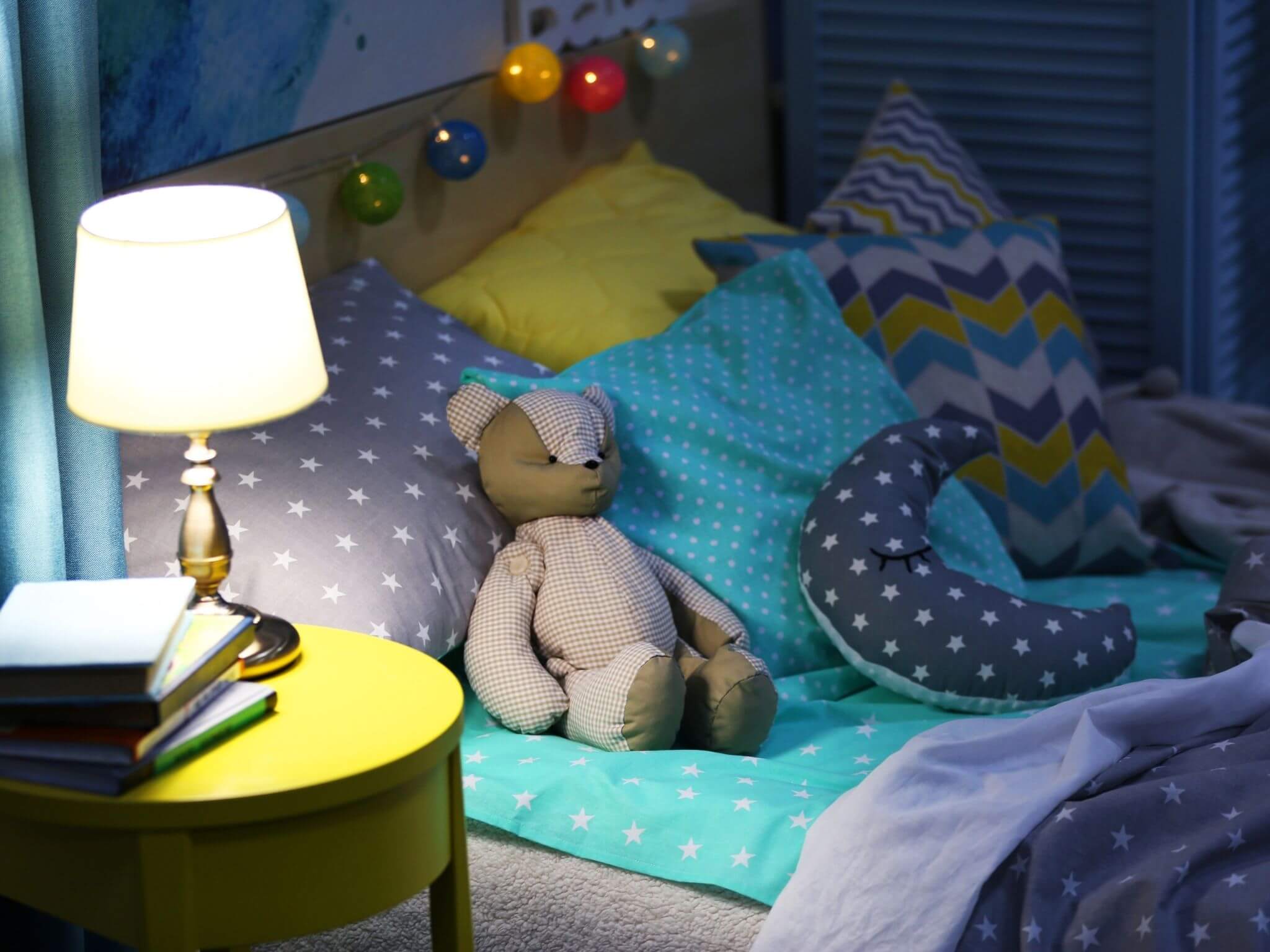 A single bed with lots of comfy children's pillows and a teddy bear and fairy lights across the headboard.