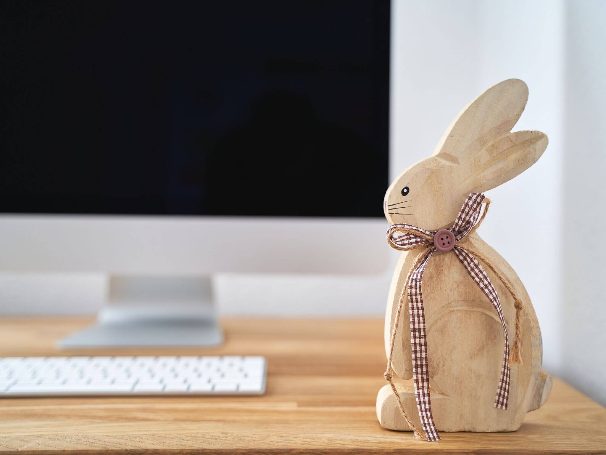A computer desk with a monitor, an Apple keyboard and a wooden carved Easter rabbit.