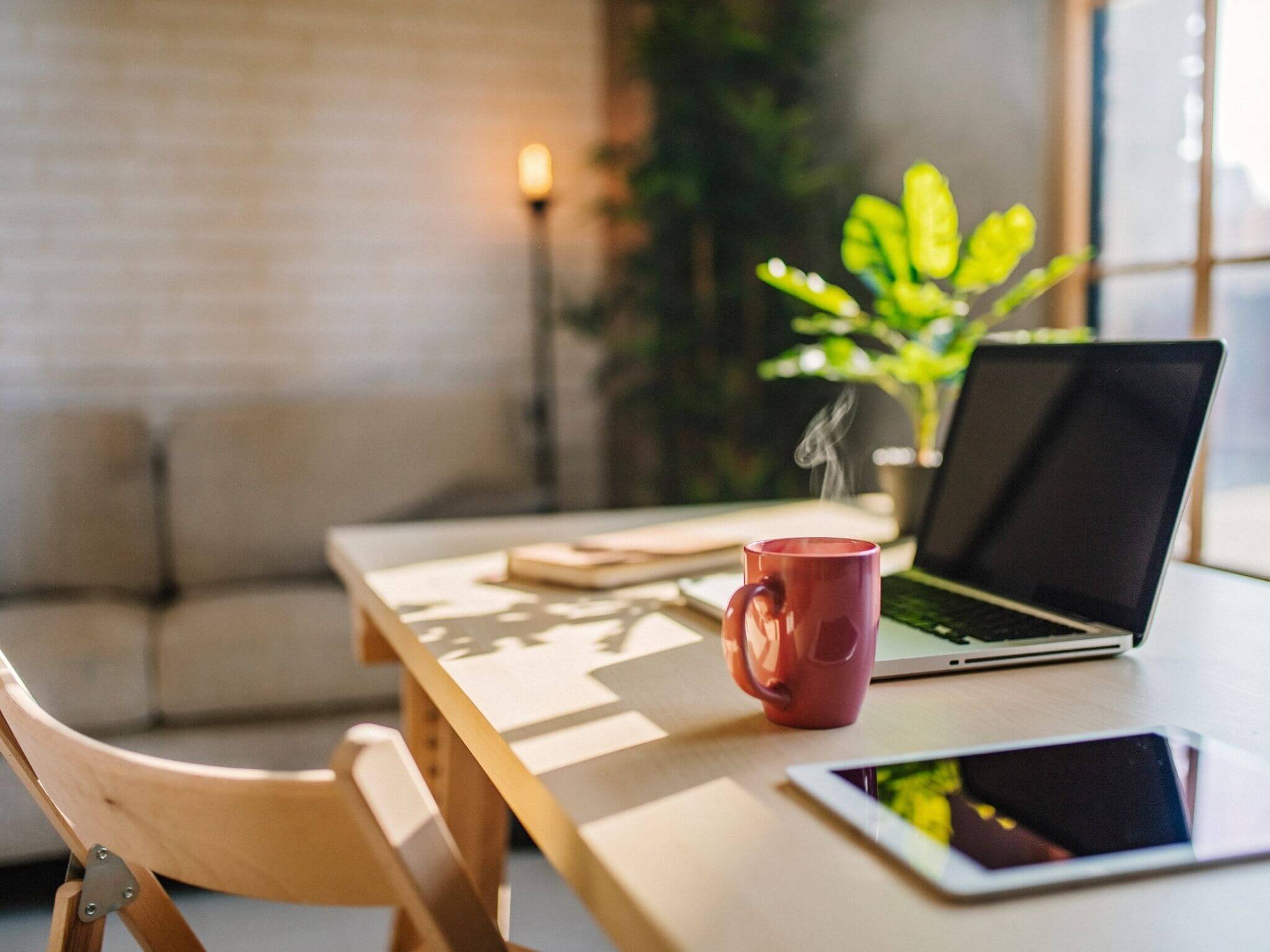 A light wood desk and chair with an open laptop on top, a red mug with a steaming coffee and an iPad. There is also a green plant in the corner of the room.