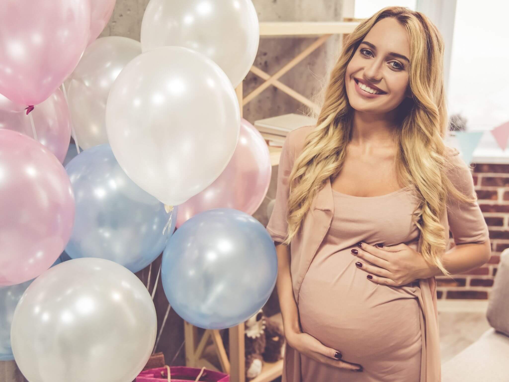 A lady in a maternity dress standing next to pink, white and blue balloons.