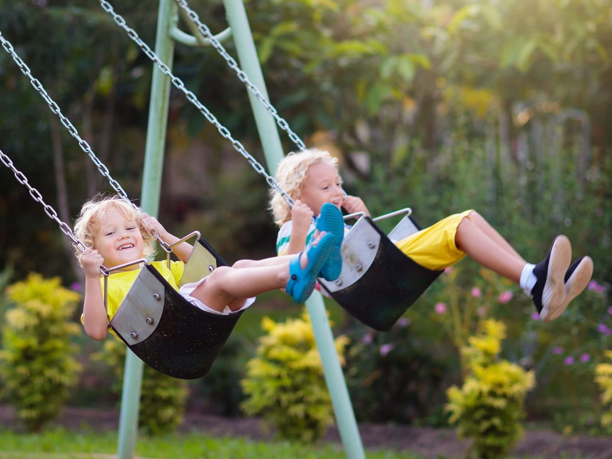 A little boy and girl both with curly blonde hair, playing on swings.