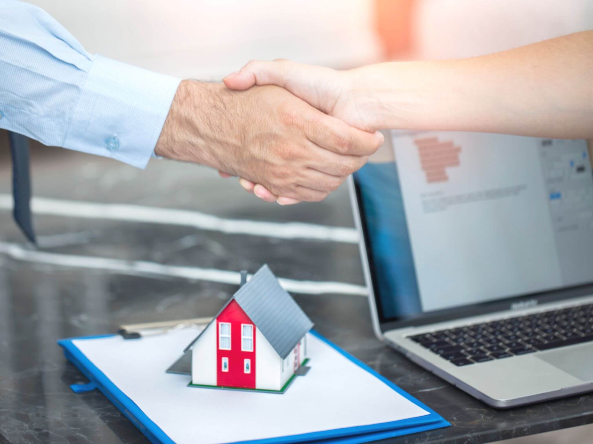 Two hands shaking across a desk with a miniature model house on top of some paperwork.