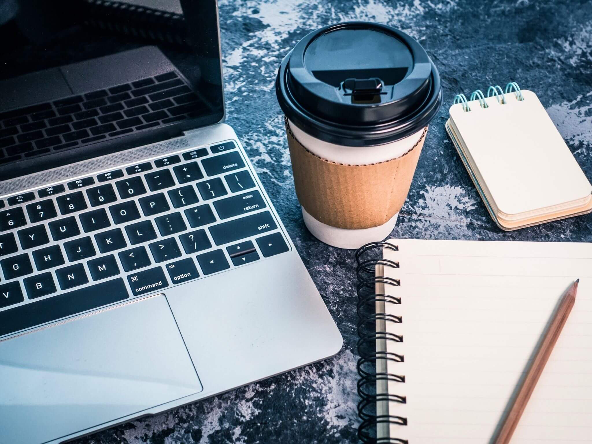 A desk with an open laptop, a travel cup and a notebook and pen on top.