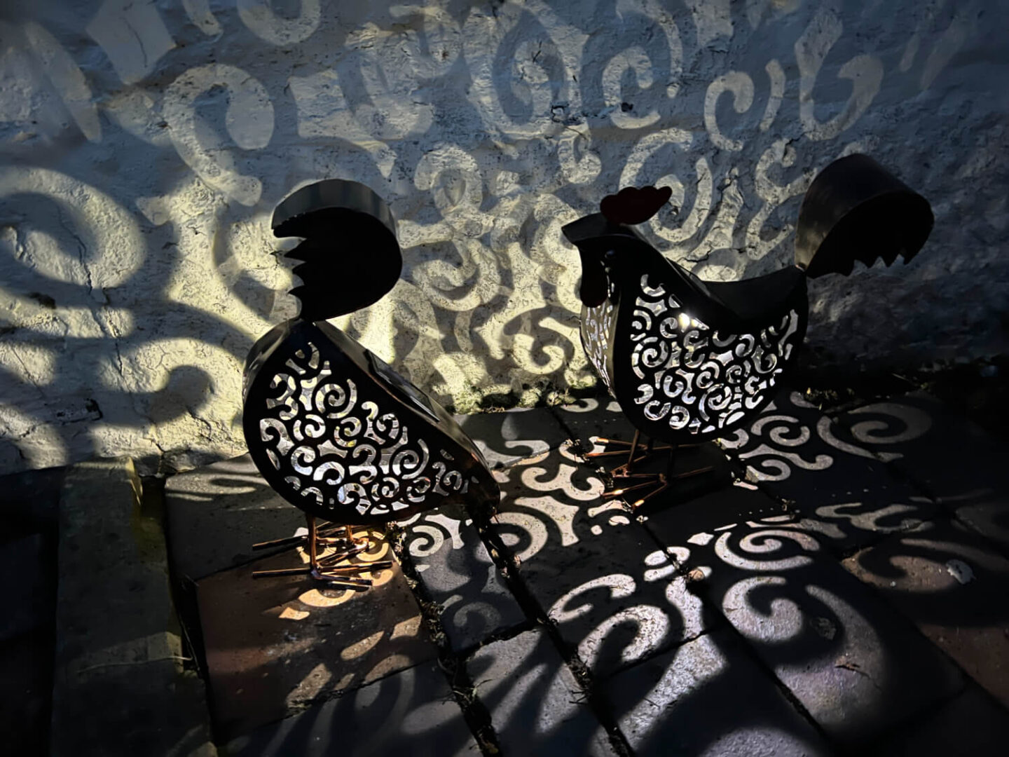 Metal framed chicken solar lights emit patterns on the wall at night.