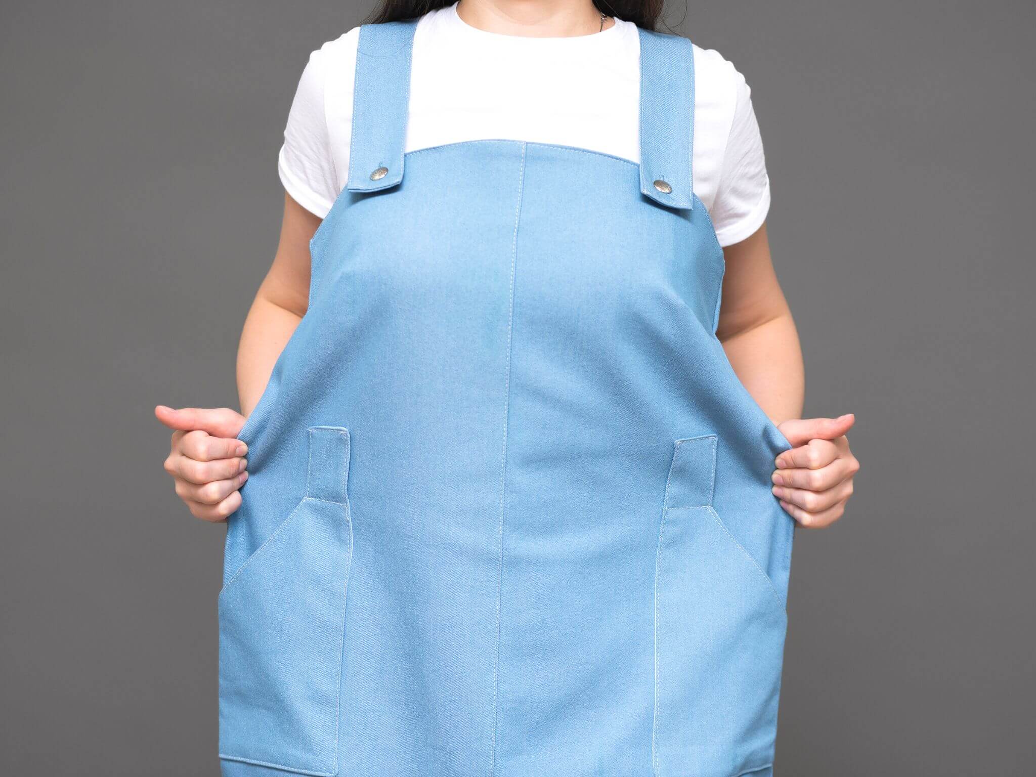 A lady with brunette hair wearing an oversized blue cotton pinafore dress. She is holding out the excess material either side of her waist.