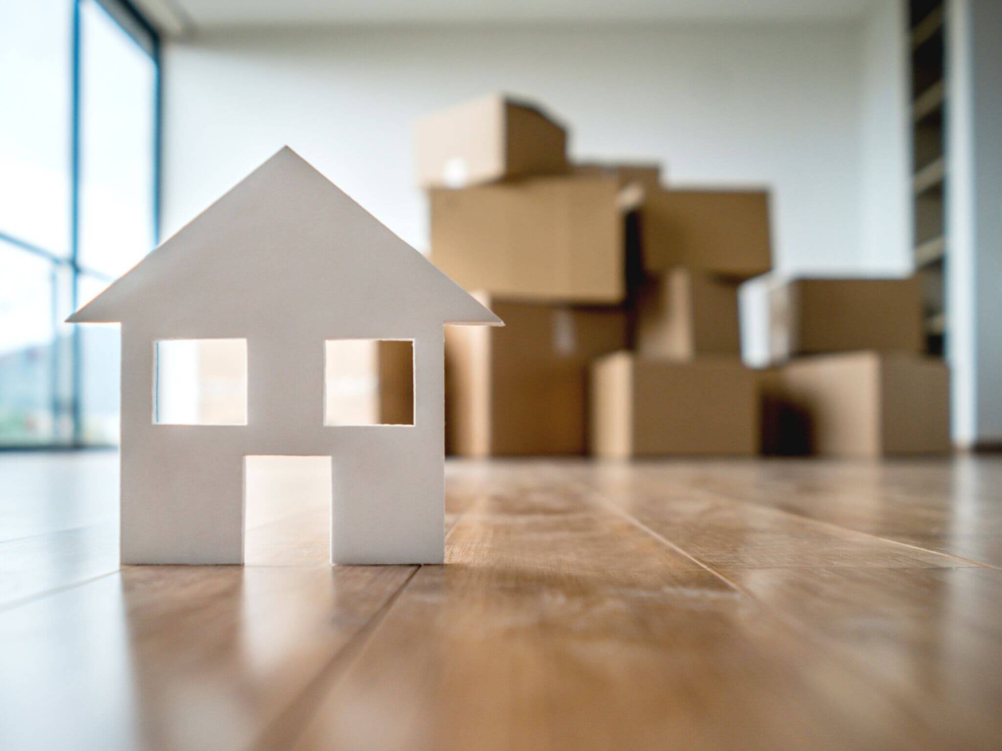 A little white miniature house on a living room floor and cardboard boxes in the background.