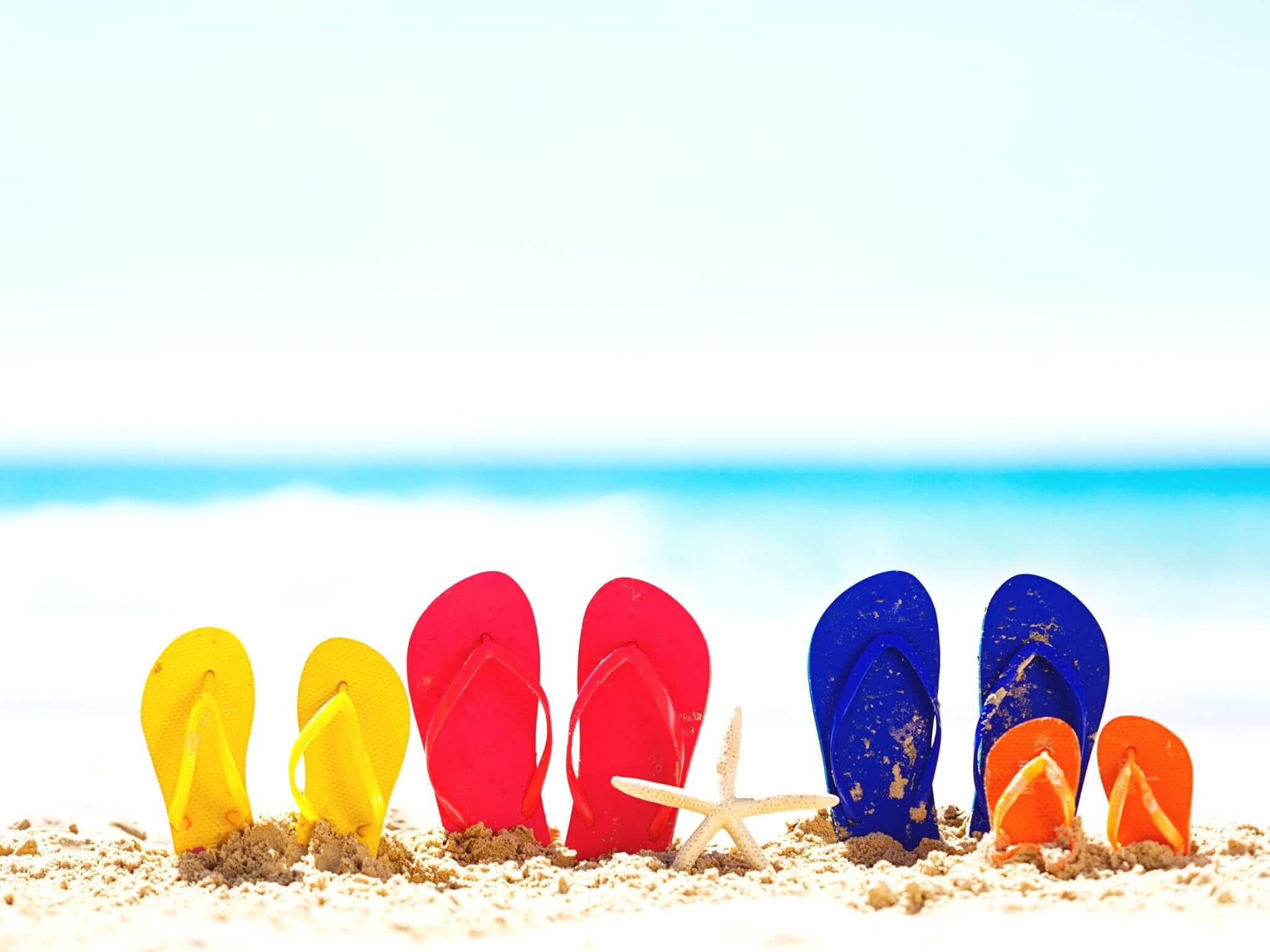 Four pairs of coloured flip-flops standing alongside each other in the sand on a sunny beach.,