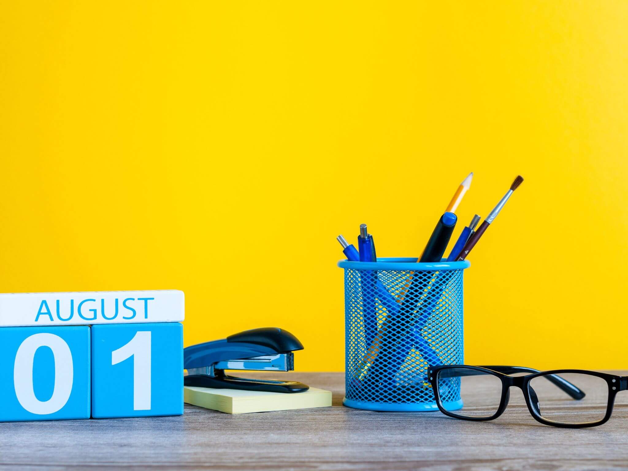 A desktop with a block calendar reading August 1st. There is also a blue pen holder, a paper stapler and a pair of black glasses.