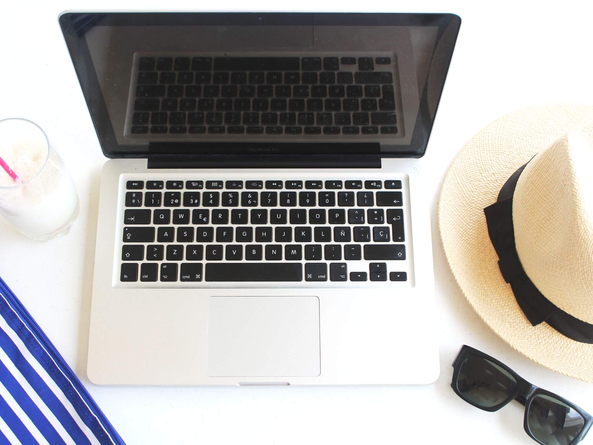 a flatlay of an open laptop on a desk with a summer straw bonnet, shades and a milkshake in a glass.