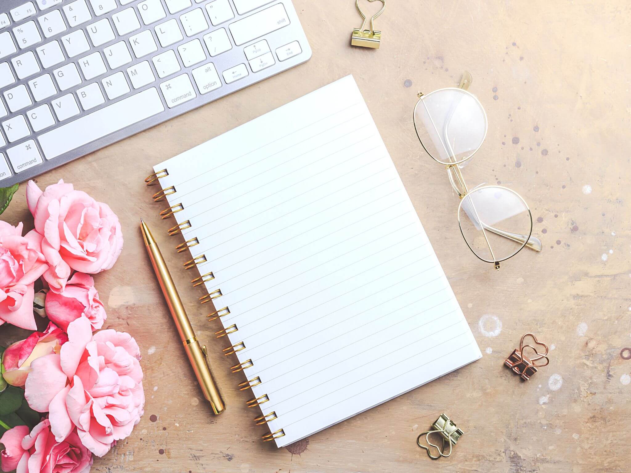 Pin peonies next to an Apple keyboard, and a notebook with a gold pen.