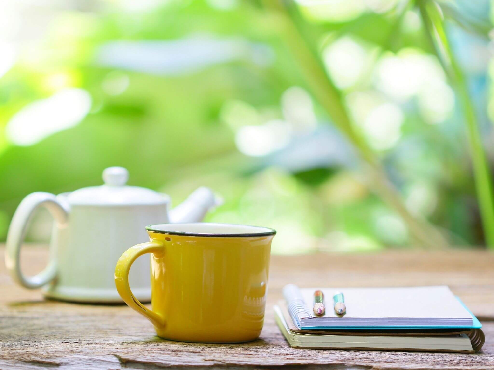A yellow enamel mug and a white teapot on a garden table with a stack of notebooks and pens to the side.