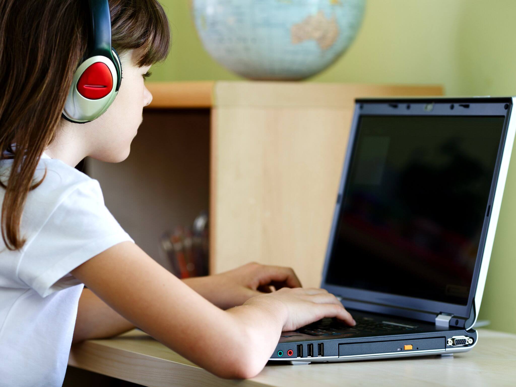 A young girl using a laptop on a desk wearing headphones.