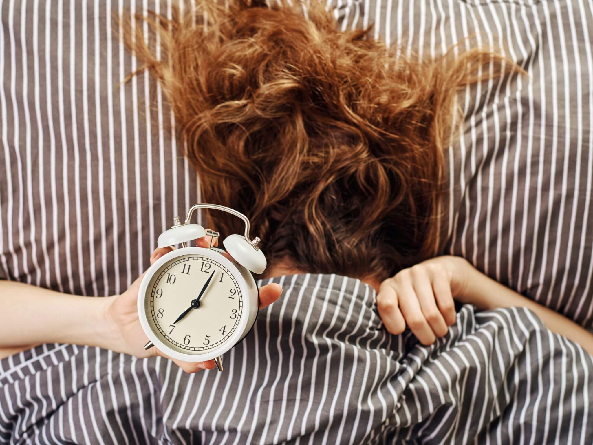A lady with long red hair, pulling the duvet over her face. There is an alarm clock to the side showing 7.05pm