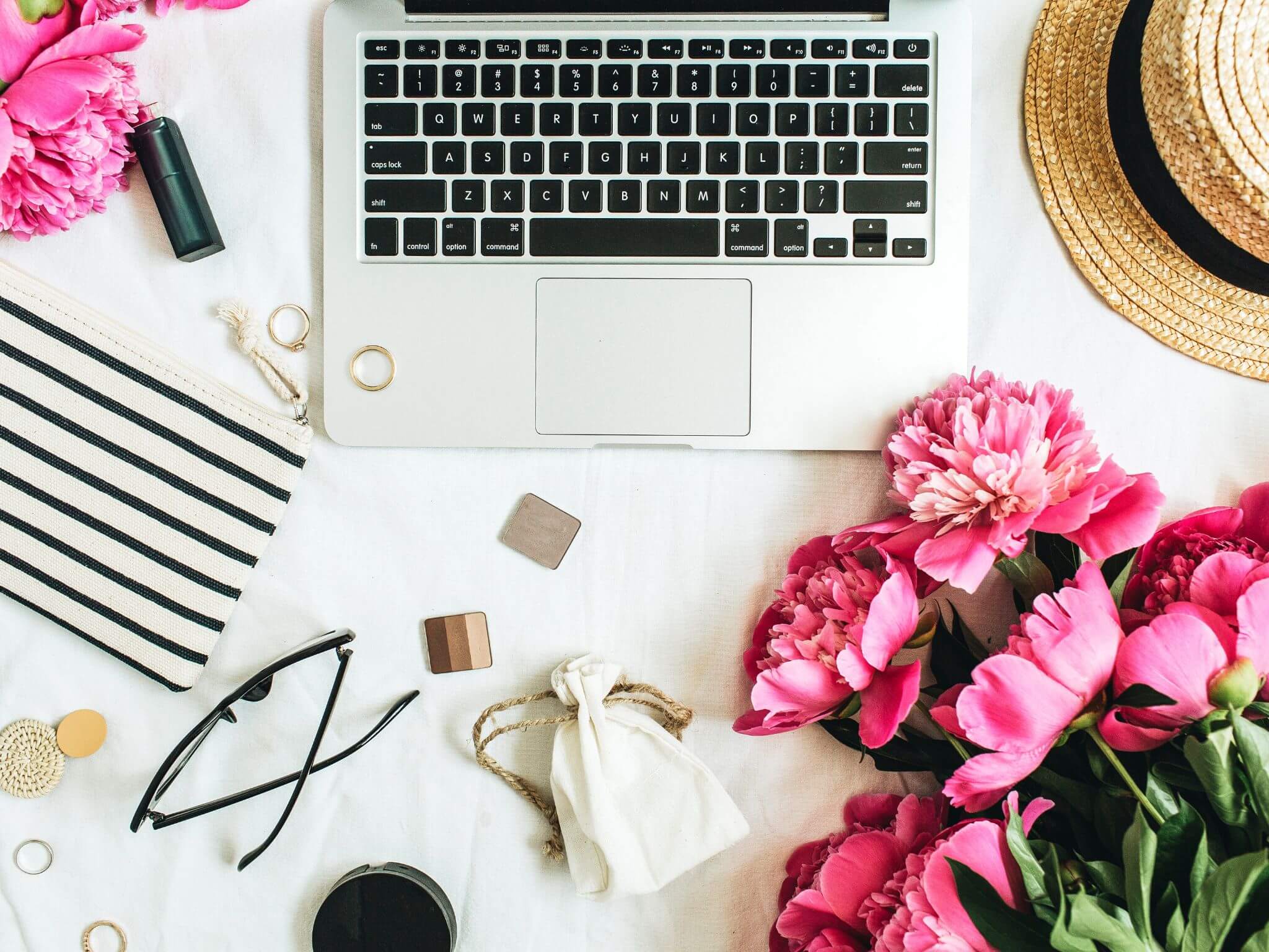 A white desk top with an open laptop, pink hydrangeas, a black and white striped notebook, a lipstick, a summer straw hat and a pair of glasses.