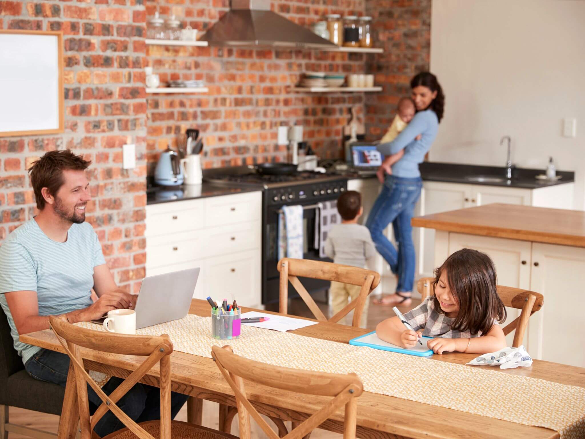 A lady holding her baby in the kitchen and her husband is sitting at the dining table with their infant daughter.