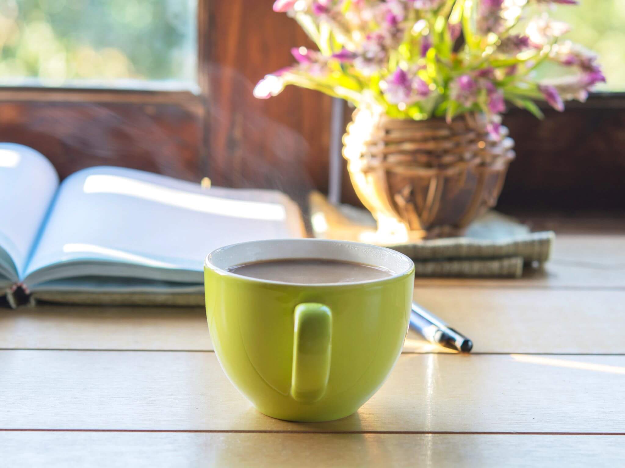 A steaming mug of coffee in a green mug. There is also an open notebook and a vase of flowers in front of the window.